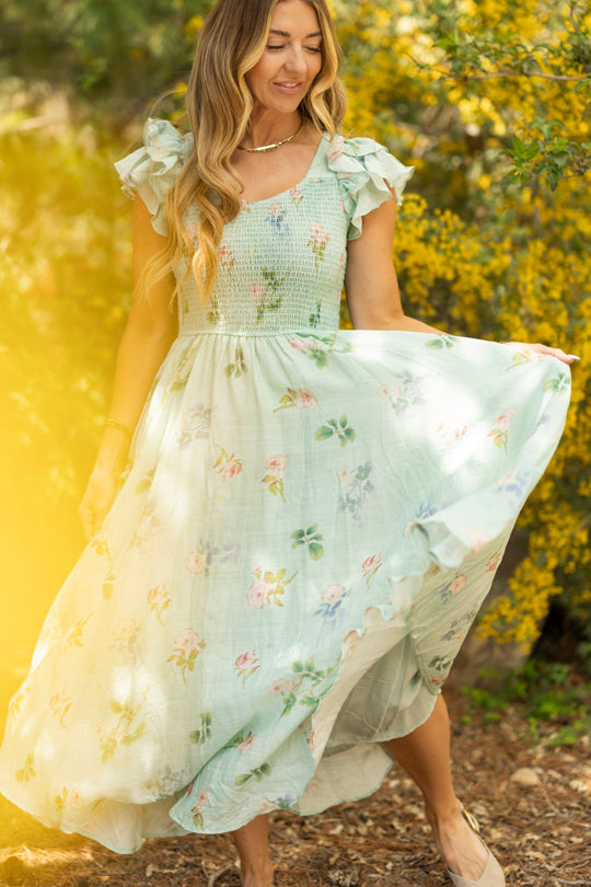 A woman in the Hattie Dress in Roselyn Blue by Ivy City Co stands outdoors, holding one side of her skirt. She has long wavy hair and is surrounded by yellow flowers and greenery, with sunlight filtering through.