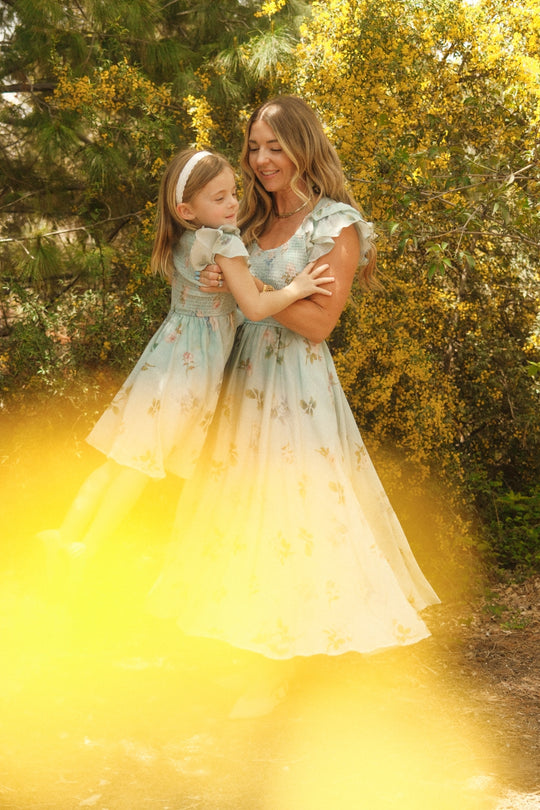 A woman and a young girl wear matching Ivy City Co Hattie Dresses in Roselyn Blue, smiling together amid yellow flowers and greenery as sunlight bathes the scene in a warm, golden glow.