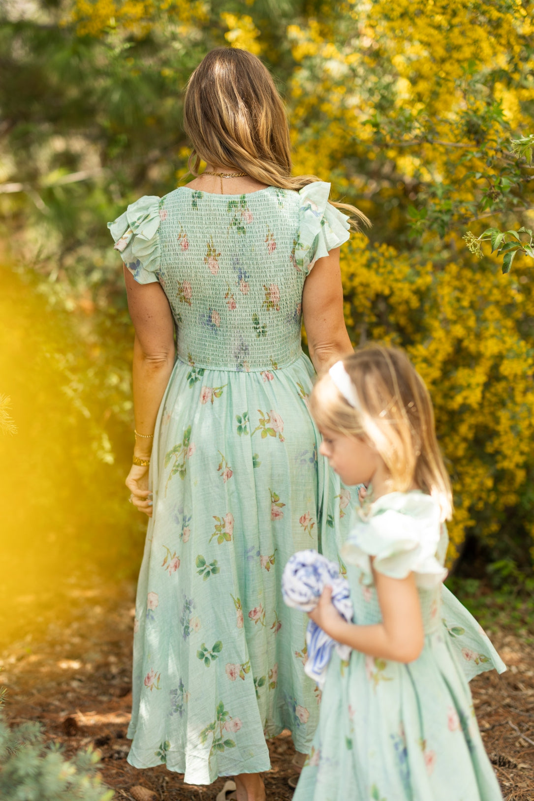 A woman and a young girl, both in matching Ivy City Co Hattie Dress in Roselyn Blue, walk outdoors among yellow flowering bushes. The woman is seen from behind while the girl, walking ahead, holds a cloth.