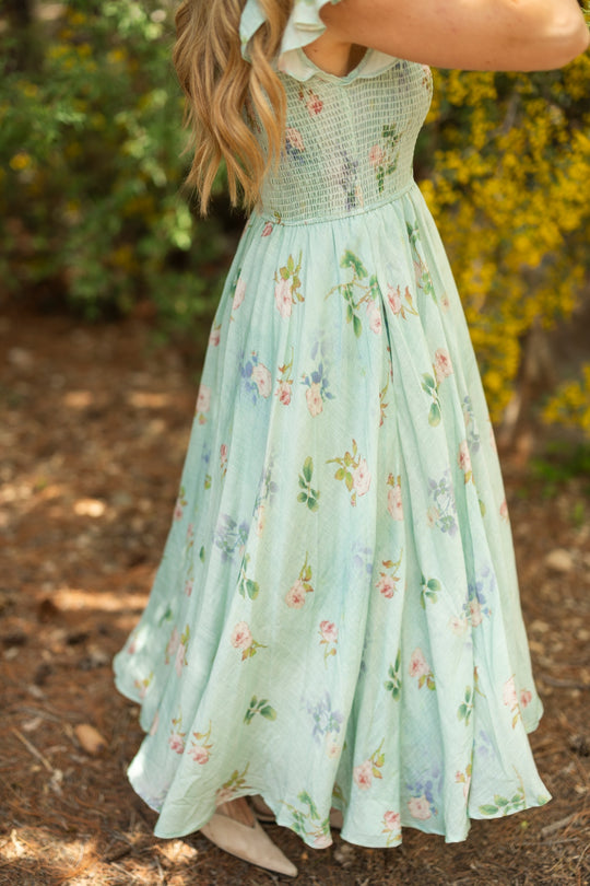 A woman stands outdoors among greenery, wearing the Ivy City Co Hattie Dress in Roselyn Blue. The sleeveless dress has a floral cotton-linen design, and her long, wavy hair is down with only her body and part of her arms visible.