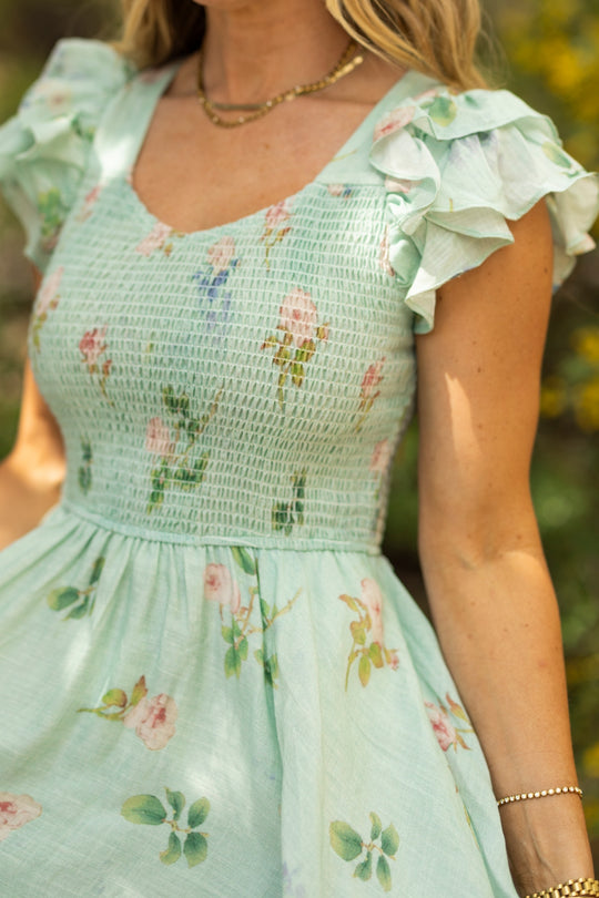 A woman, shown outdoors from the waist up, wears the Hattie Dress in Roselyn Blue by Ivy City Co—an A-line cotton-linen midi dress with a smocked bodice and ruffled sleeves—paired with gold jewelry.