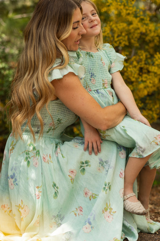 A woman and a young girl wear matching Ivy City Co Hattie Dresses in Roselyn Blue as they sit outdoors, sharing a sweet moment surrounded by greenery and yellow flowers.