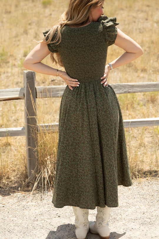 Outdoors by a wooden fence, a woman stands with her hands on her lower back, facing away and wearing the Ivy City Co Hattie Dress in Woodland Floral—a green floral dress featuring a smocked bodice and ruffled sleeves—paired with white cowboy boots. Maternity friendly and effortlessly stylish, she is framed by dry grass and a dirt path.