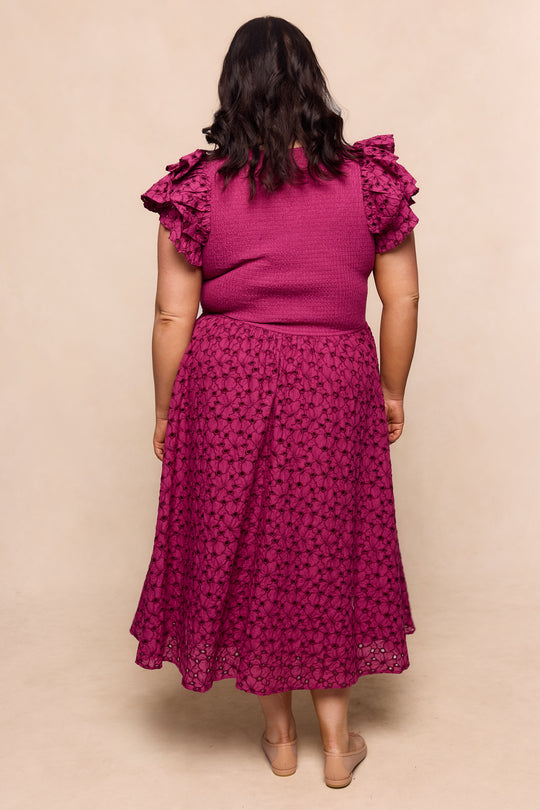 A woman with medium-length dark hair stands barefoot, facing away against a beige background, wearing the Ivy City Co Hattie Dress in Magenta—a sleeveless embroidered dress with ruffled shoulders and a matching lace midi skirt.