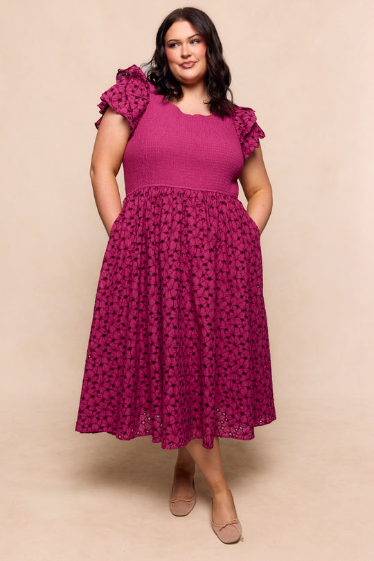 A woman wears the Ivy City Co Hattie Dress in Magenta, featuring embroidery, textured short sleeves, ruffled shoulders, and a floral lace midi skirt. She stands before a beige backdrop with loose dark hair and nude flats.