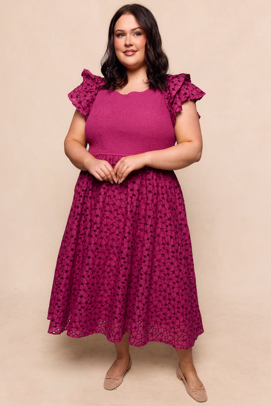 A woman with dark hair smiles, standing forward in light flats, wearing Ivy City Co's Hattie Dress in Magenta—a midi-length dress with embroidery, short sleeves, ruffled shoulders, and a textured skirt—against a neutral backdrop.