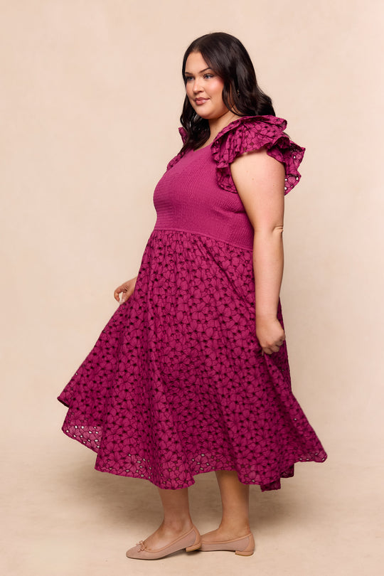 A woman with long dark hair wears the Hattie Dress in Magenta by Ivy City Co, featuring embroidery, ruffled sleeves, and a textured pattern. She stands sideways, smiling and holding the skirt, styled with nude flats against a beige background.