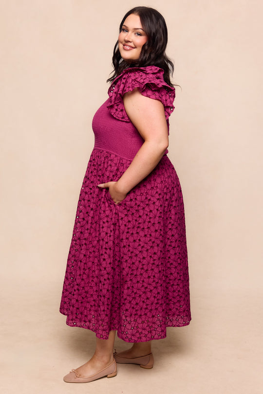 A woman smiles sideways, wearing the Ivy City Co Hattie Dress in Magenta—a sleeveless, maternity-friendly, midi-length dress with ruffled shoulders and a lace skirt. She pairs it with nude flats, one hand in her pocket against a neutral background.