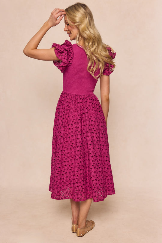 A woman with long blonde hair stands facing away, wearing the Hattie Dress in Magenta by Ivy City Co—a chic, maternity-friendly, embroidered midi dress with ruffled sleeves and eyelet details—paired with flat sandals. She raises one hand near her head.
