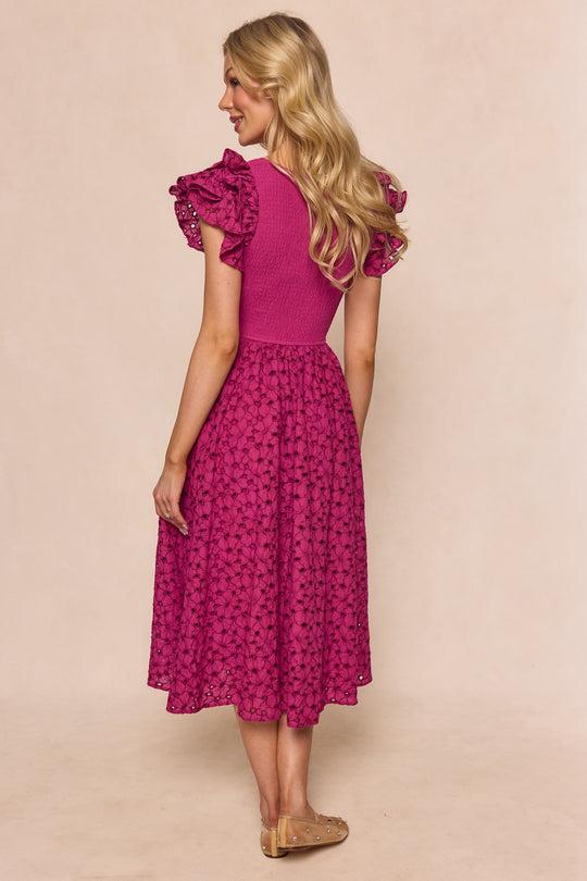 A woman with long blonde hair stands on a light background wearing the Ivy City Co Hattie Dress in Magenta, a midi-length style with short ruffled sleeves and a textured embroidered floral pattern, paired with beige flats.