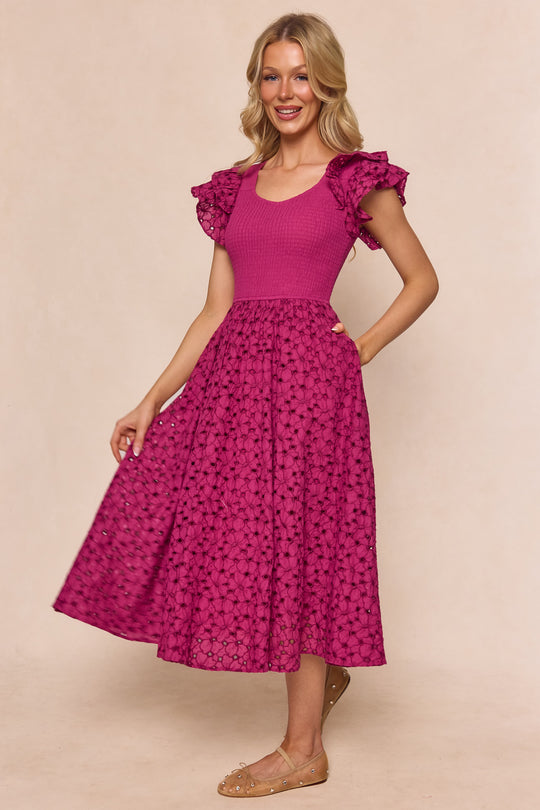 A woman wears the Ivy City Co Hattie Dress in Magenta—a maternity-friendly, short-sleeve midi dress with ruffled shoulders and a patterned skirt—smiling with one hand on her hip, against a light beige background.