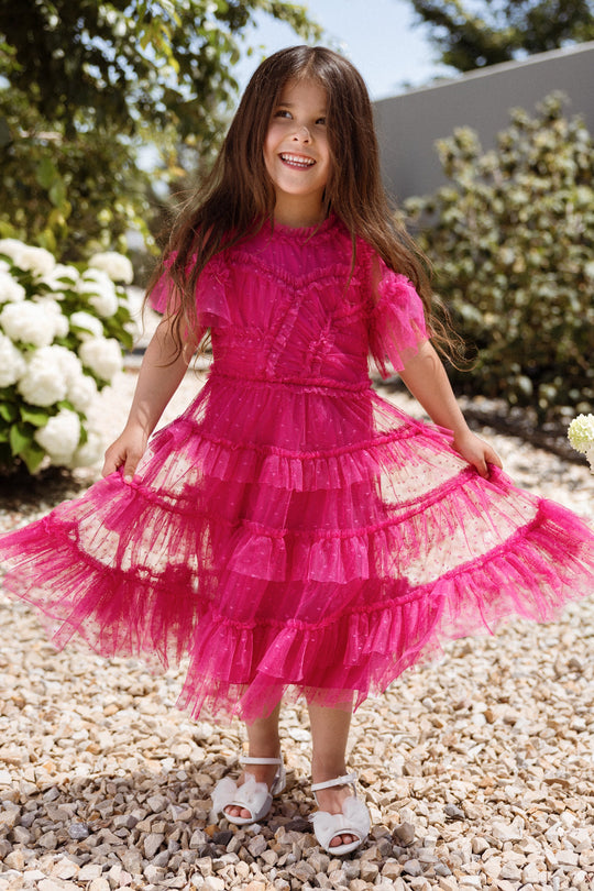 A young girl stands outdoors on gravel, beaming as she holds out her Ivy City Co Mini Whimsical Dress in Fuchsia, which features a layered tulle skirt. She pairs the dress with white shoes, while lush green bushes with white flowers provide a sunny backdrop.