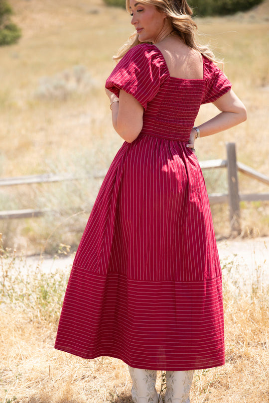 A woman stands outdoors on dry grass, wearing the Ivy City Co Julie Dress in Cranberry Pinstripe with subtle puff sleeves and white boots, facing away while looking over her shoulder on a sunny day.