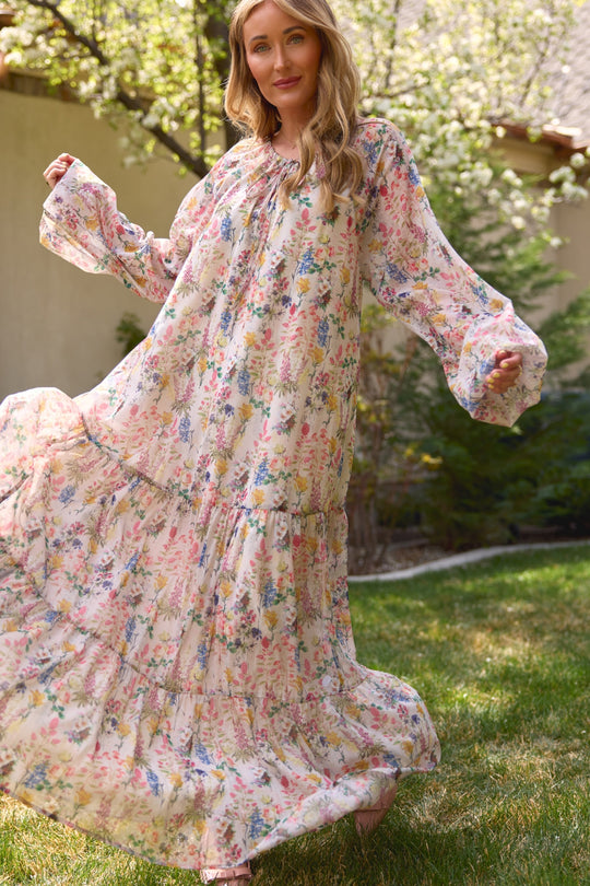 A woman smiles outdoors on grass, holding out the Ivy City Co Imogen Dress in Cordelia Floral—a flowy, long sleeve, maternity friendly maxi. Greenery and a house appear in the background.