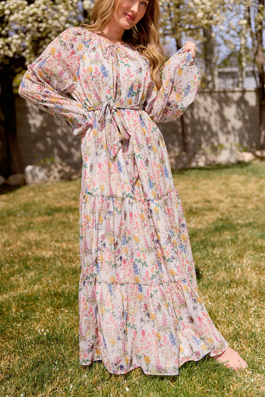 A woman stands barefoot on grass in the Ivy City Co Imogen Dress in Cordelia Floral—a flowy chiffon maxi with billowy sleeves and tied waist—surrounded by blooming trees for an effortlessly elegant, maternity-friendly look.