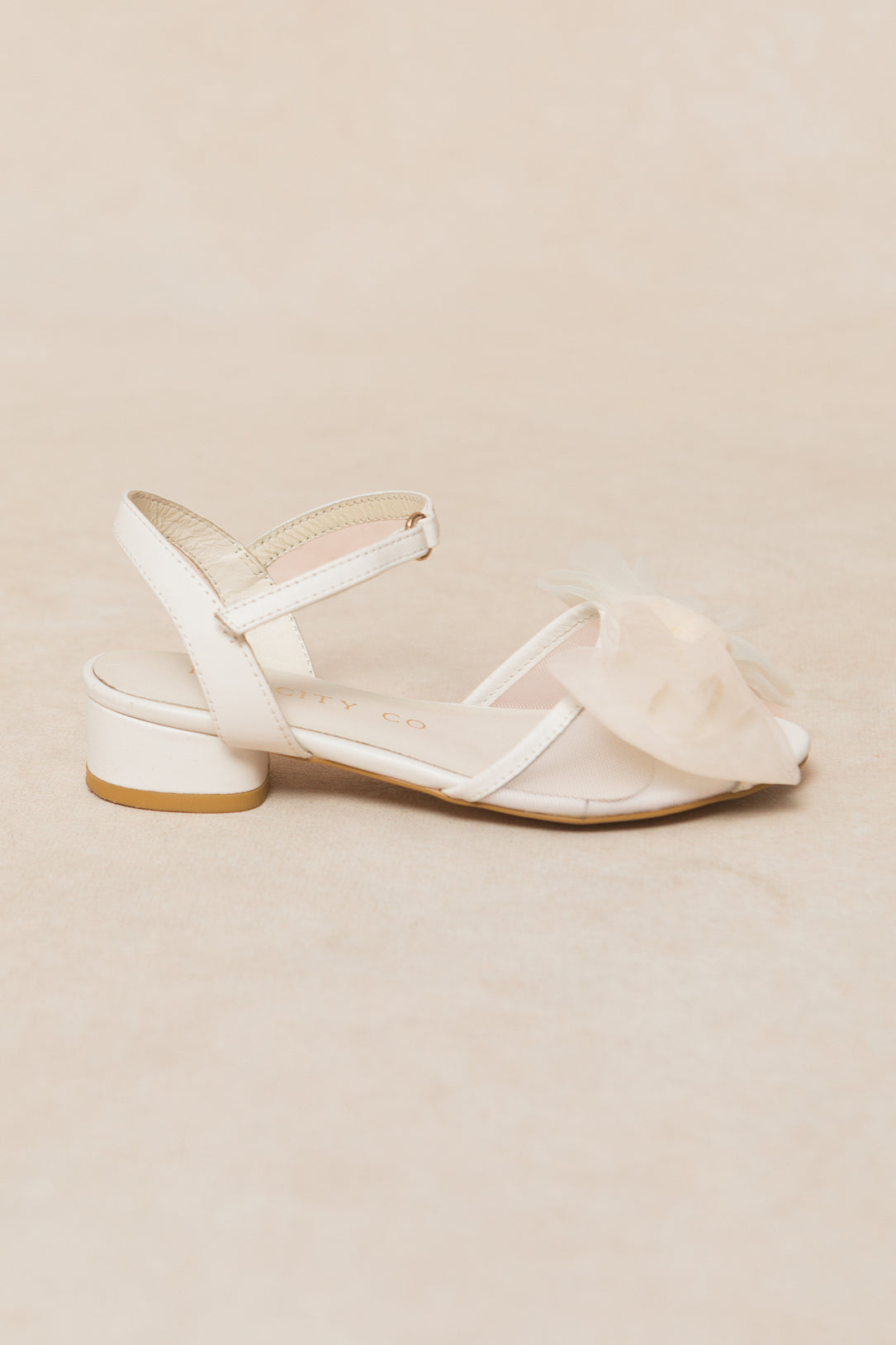 The Ivy City Co Mini Ivy Bow Heel in White, featuring a low block heel, ankle strap, and a large sheer tulle bow on the front, is displayed against a light beige background.