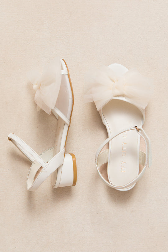 The Mini Ivy Bow Heel in White by Ivy City Co features open toes, low block heels, ankle straps, and oversized sheer tulle bows on the front straps, shown against a neutral beige background.