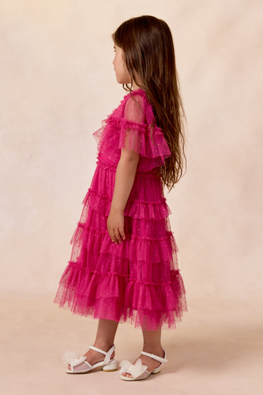 A young girl with long brown hair stands sideways against a beige background, wearing the Ivy City Co Mini Whimsical Dress in Fuchsia, which features flutter sleeves and a layered tulle skirt. She completes her look with white sandals.