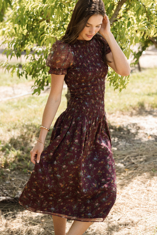 A woman wearing the Ivy City Co Jennifer Dress in Aubergine Elysian Vine, featuring a smocked bodice and maternity-friendly design, stands outdoors near lush green trees on a sunny day, gently touching her hair and looking down in a natural setting.