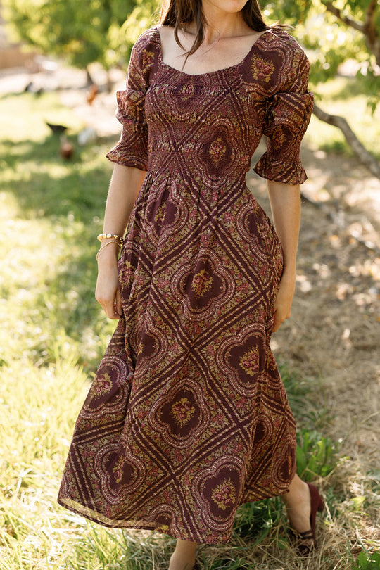 A woman wearing the Josephine Dress in Nocturne Diamond Rose by Ivy City Co, featuring a burgundy floral print with puff sleeves, a smocked bodice, and a square neckline, stands outdoors in a garden setting.