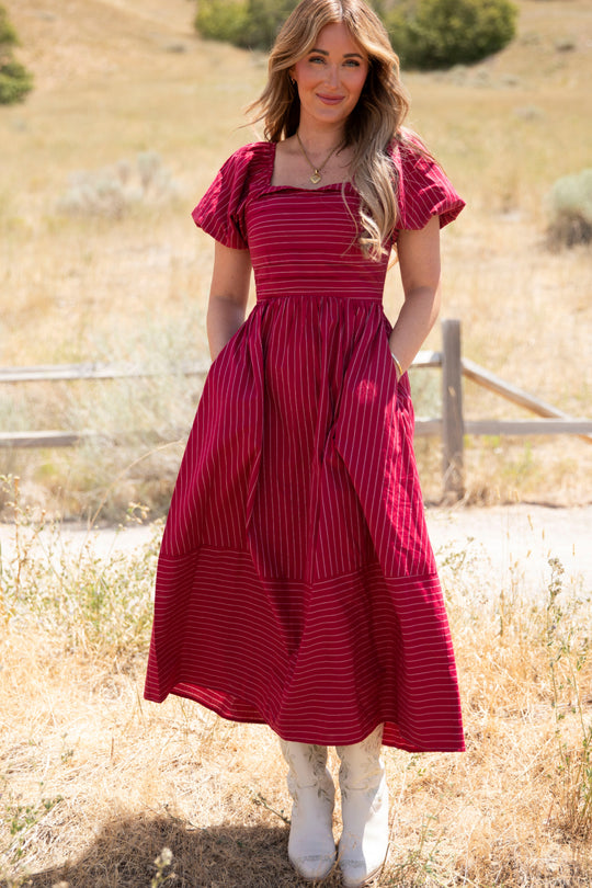 A woman stands outdoors in a sunny field wearing the Ivy City Co Julie Dress in Cranberry Pinstripe, featuring flutter sleeves, a square neckline, smocked bodice, and pockets; she pairs it with white cowboy boots.