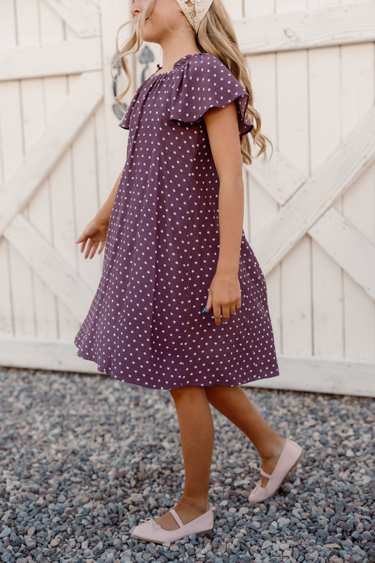 A young girl in the Ivy City Co Teen Blakely Dress in Aubergine Ditsy Dot, featuring flutter sleeves, pairs it with pink shoes as she walks on gravel before a white wooden gate, her long hair partly adorned with a lace headband.
