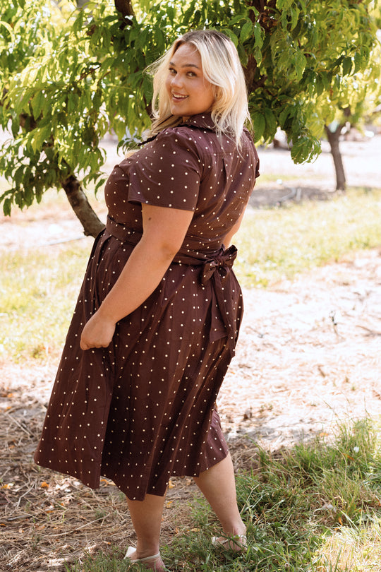 A woman wears the Ivy City Co Kit Dress in Warm Cocoa Dainty Dot, standing outdoors near green trees and smiling over her shoulder.