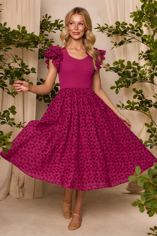 A woman smiles indoors among green plants, wearing the Ivy City Co Hattie Dress in Magenta—a midi-length, embroidered dress with ruffled sleeves and a flared skirt. She holds out her skirt and stands in sandals on a beige floor.