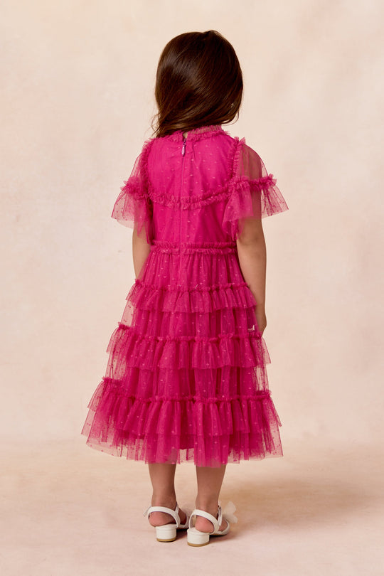 A young girl with long brown hair stands facing away, wearing the Ivy City Co Mini Whimsical Dress in Fuchsia with flutter sleeves and a layered tulle skirt, paired with white sandals against a light beige background.