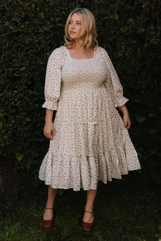 A blonde woman stands outdoors before green bushes, wearing the Ivy City Co Madeline Dress in Blue—a floral, smocked midi that's maternity and nursing friendly—paired with brown platform shoes. She gazes calmly to the side.