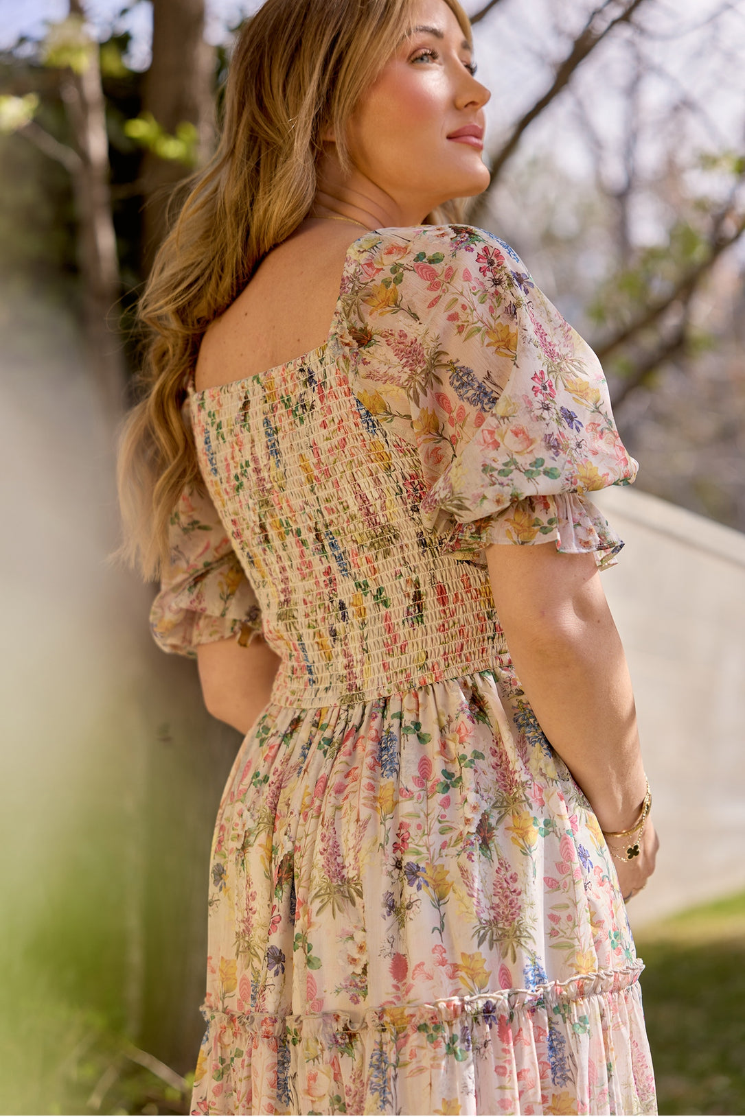 A woman stands outdoors in Ivy City Co’s Madeline Dress in Cordelia Floral, featuring puffed short sleeves and a smocked bodice. Sunlight highlights her long, wavy hair as she looks to the side, surrounded by greenery and trees.