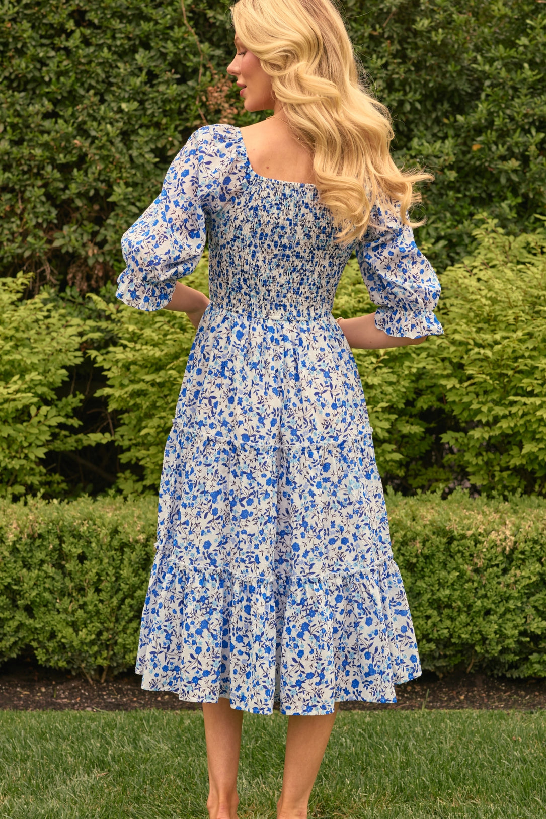 A woman with long blonde hair stands on grass, facing away, wearing the Ivy City Co Madeline Dress in Ditsy Cobalt Floral—a blue and white cotton midi that’s maternity and nursing friendly—amid green bushes and trees.