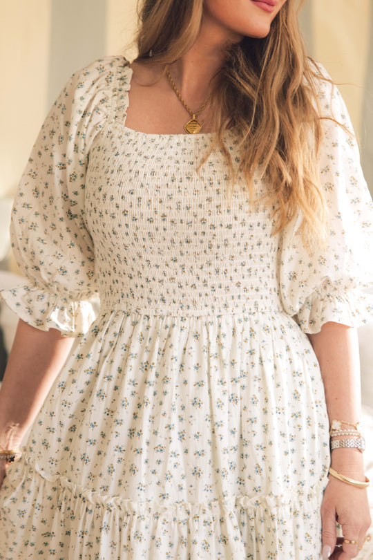 A woman models the Ivy City Co Madeline Dress in Blue, a maternity and nursing-friendly midi with a smocked bodice and puffed sleeves, styled with gold jewelry. Soft lighting showcases her long hair and the dress’s elegant floral design.