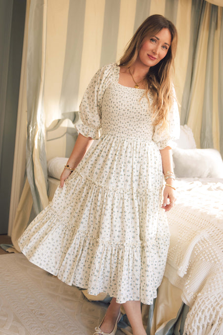 A woman with long hair wears the Ivy City Co Madeline Dress in Blue, a floral maternity nursing-friendly midi with a smocked bodice, standing by a bed in an elegantly lit bedroom with light decor.