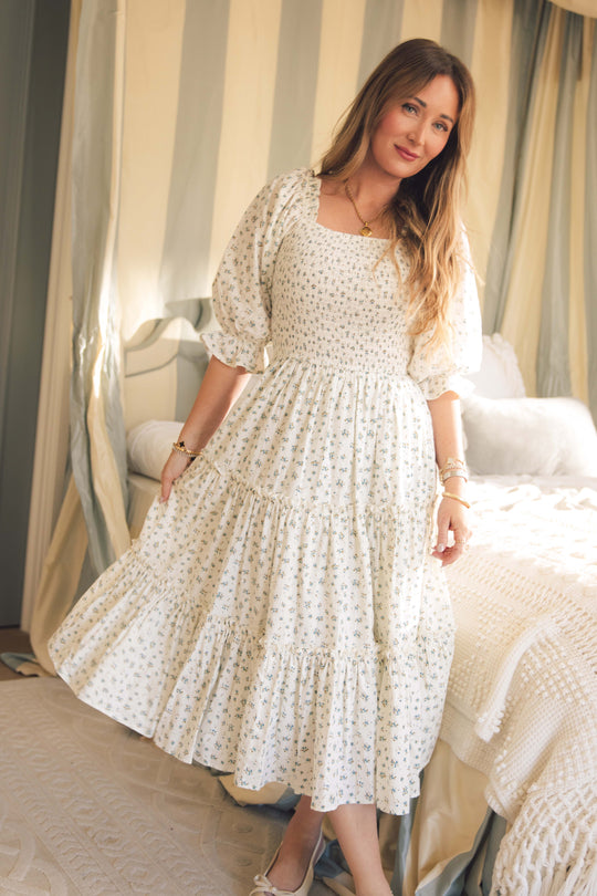 A woman with long hair wears the Ivy City Co Madeline Dress in Blue, a floral maternity nursing-friendly midi with a smocked bodice, standing by a bed in an elegantly lit bedroom with light decor.