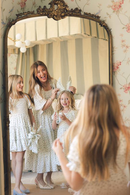 A woman wears the Ivy City Co Madeline Dress in Blue, smiling with two young girls in matching outfits by an antique mirror in a vintage-style room, holding playful bunny ear headbands.