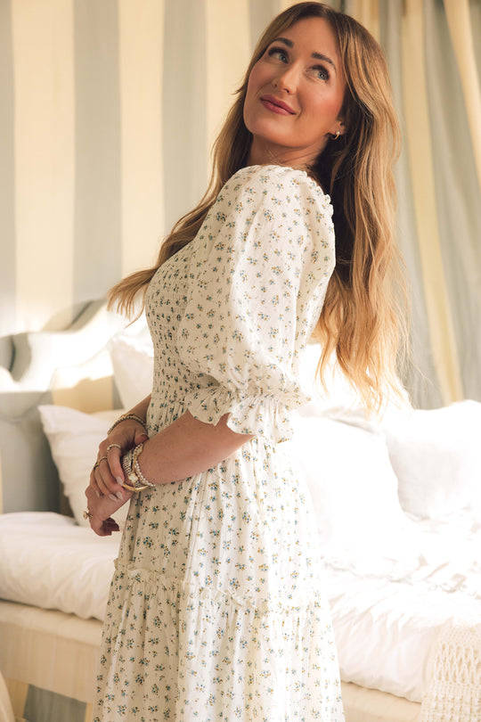 A woman with long, wavy blonde hair models the Ivy City Co Madeline Dress in Blue—a floral midi dress with puffed sleeves and a smocked bodice. She smiles softly indoors, highlighted by gentle sunlight. Maternity and nursing friendly style.