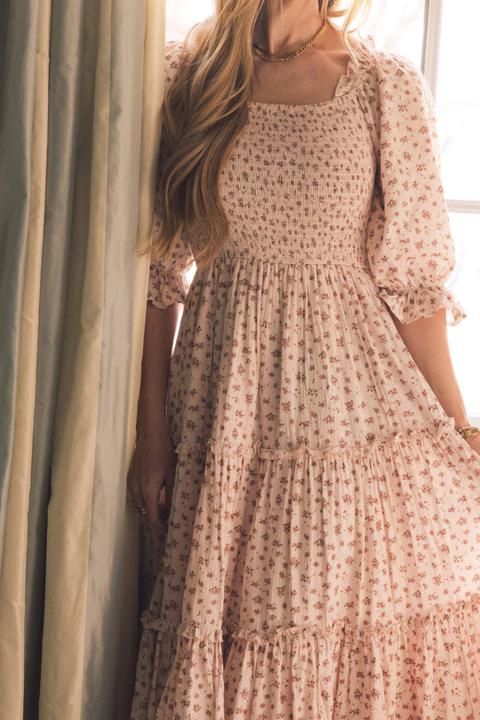 A woman in the Ivy City Co Madeline Dress In Pink—a long, floral dress with puffed sleeves—stands by a window with light curtains. She has wavy hair over one shoulder and wears bracelets and a necklace. Maternity friendly.