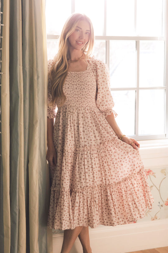 A woman stands by a window, smiling and holding her Ivy City Co Madeline Dress In Pink—a long, floral dress with ruffled tiers and puffed sleeves that's both elegant and maternity friendly. Soft light creates a warm, gentle atmosphere.