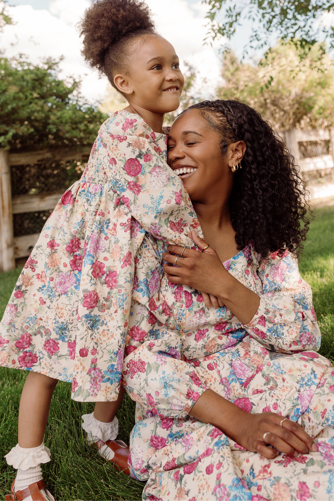 A smiling woman hugs a cheerful young girl as they sit on green grass outdoors, with trees and a fence in the background. Both wear matching Ivy City Co Mini Adelpha Dresses in Autumn Amity Floral, featuring smocked bodices and playful balloon sleeves.