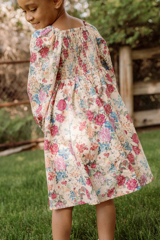 A young child in the Ivy City Co Mini Adelpha Dress in Autumn Amity Floral with charming balloon sleeves stands on the grass, looking down and smiling. The smocked bodice adds a delicate touch, while a wooden fence and green trees are visible in the background.