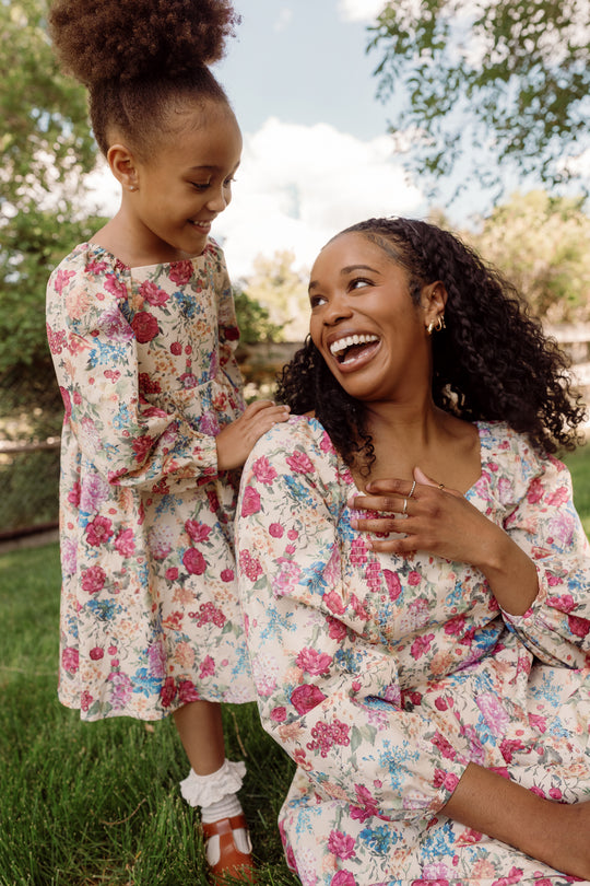 A smiling woman and young girl, both dressed in matching Ivy City Co Mini Adelpha Dresses in Autumn Amity Floral with smocked bodices, sit together on green grass outdoors. The woman looks up at the girl, and they share a joyful moment.