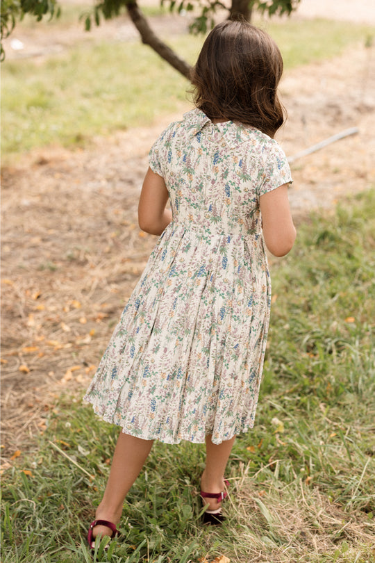 A young girl wearing the Ivy City Co Mini Amber Dress in Autumn Bouquet and sandals walks away on a grassy path outdoors, with trees in the background.