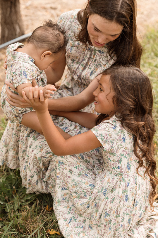 A woman and two children in matching floral dresses sit on the grass. The older child, dressed in Ivy City Co’s Mini Amber Dress in Autumn Bouquet, gently holds the baby’s arm while the woman, wearing a flowing midi-length dress, looks down lovingly. They share a warm, tender moment outdoors.