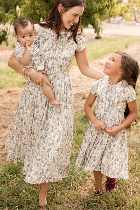 A woman holding a baby and walking with a young girl, all smiling in matching Ivy City Co dresses—including the delightful Mini Amber Dress in Autumn Bouquet—as they enjoy time outdoors.