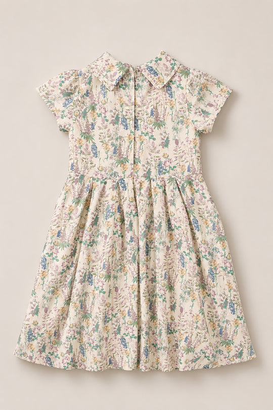 The Mini Amber Dress in Autumn Bouquet by Ivy City Co is a short-sleeved, collared viscose dress for children, adorned with a pastel floral pattern. It features buttons down the front and a gathered skirt, shown laid flat on a neutral background.