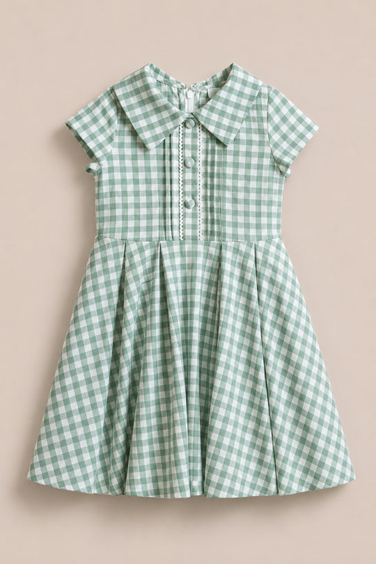 The Ivy City Co Mini Amber Dress in Sage Gingham features short sleeves, a wide collar, front buttons, decorative trim, and a pleated skirt. This knee-length cotton midi dress is shown against a plain beige background.