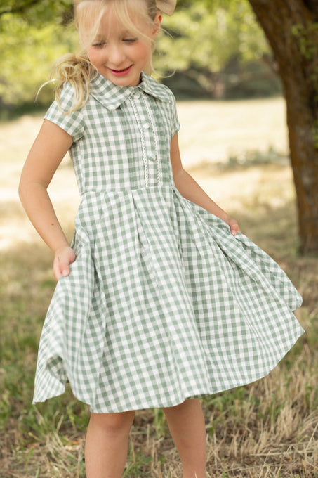 A young girl wears the Ivy City Co Mini Amber Dress in Sage Gingham, smiling as she gently holds out her pleated skirt while standing on grass with trees and sunlight in the background.