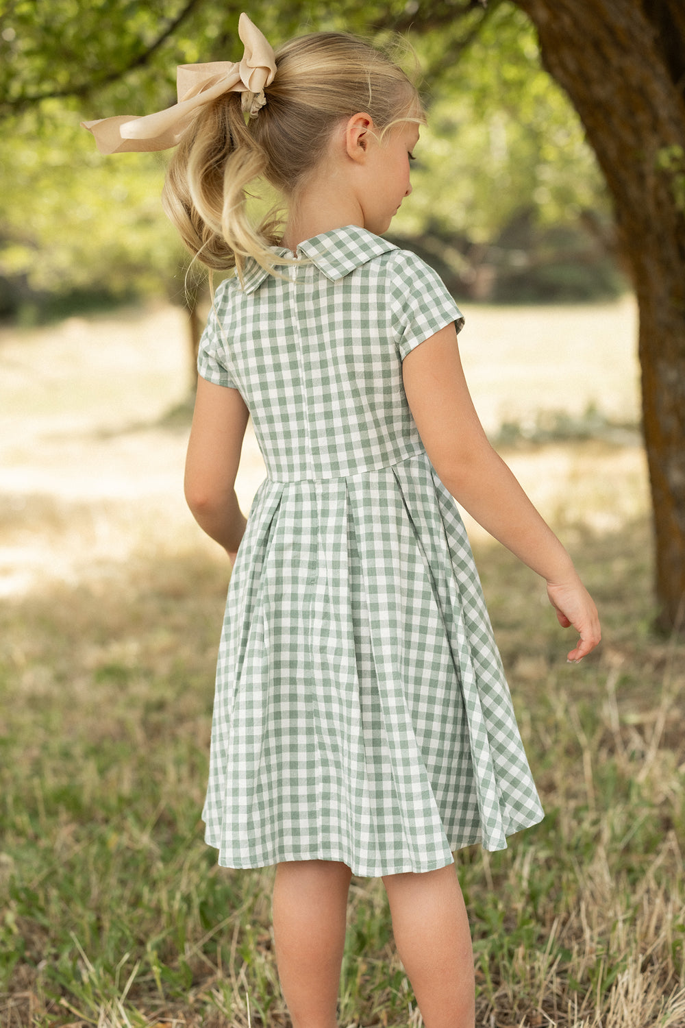 A young girl with blonde hair in a large beige bow stands outdoors near a tree, wearing the Ivy City Co Mini Amber Dress in Sage Gingham, featuring a pleated skirt. She is on grass with softly blurred greenery in the background.