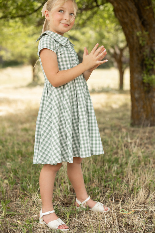 A young girl with blonde hair in a ponytail smiles and claps her hands while wearing the Ivy City Co Mini Amber Dress in Sage Gingham and white sandals, standing on grass near a tree.